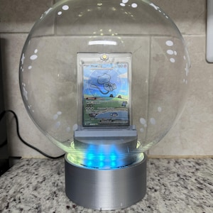 Op de afbeelding: Een heldere, bolvormige displaycase met een Pokémon-verzamelkaart erin. De kaart toont een blauw Mew-personage. De basis is zilverkleurig met blauwe LED-verlichting. De bovenkant van de bol heeft witte decoratieve stippen.