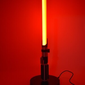Op de afbeelding: Een rode lightsaber-lamp met een zwarte voet en een snoer. De lightsaber is aan en gloeit.