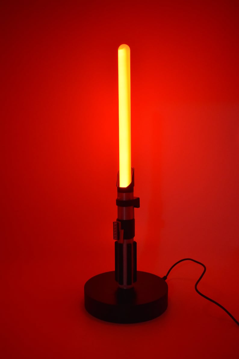 Op de afbeelding: Een rode lightsaber-lamp met een zwarte voet en een snoer. De lightsaber is aan en gloeit.