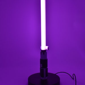 K&ouml;nnte beinhalten: Eine lila Lichtschwert-Lampe mit einer leuchtenden wei&szlig;en Klinge. Die Lampe hat einen schwarzen Sockel und Griff mit silbernen und schwarzen Details. Der Hintergrund ist einfarbig lila.