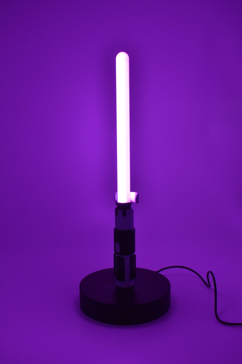 K&ouml;nnte beinhalten: Eine lila Lichtschwert-Lampe mit einer leuchtenden wei&szlig;en Klinge. Die Lampe hat einen schwarzen Sockel und Griff mit silbernen und schwarzen Details. Der Hintergrund ist einfarbig lila.