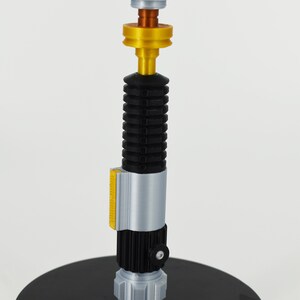 K&ouml;nnte beinhalten: Eine silberne und schwarze Lichtschwert-Replik mit einem goldenen Akzentringe und einer wei&szlig;en Spitze. Das Lichtschwert steht auf einem schwarzen Sockel.