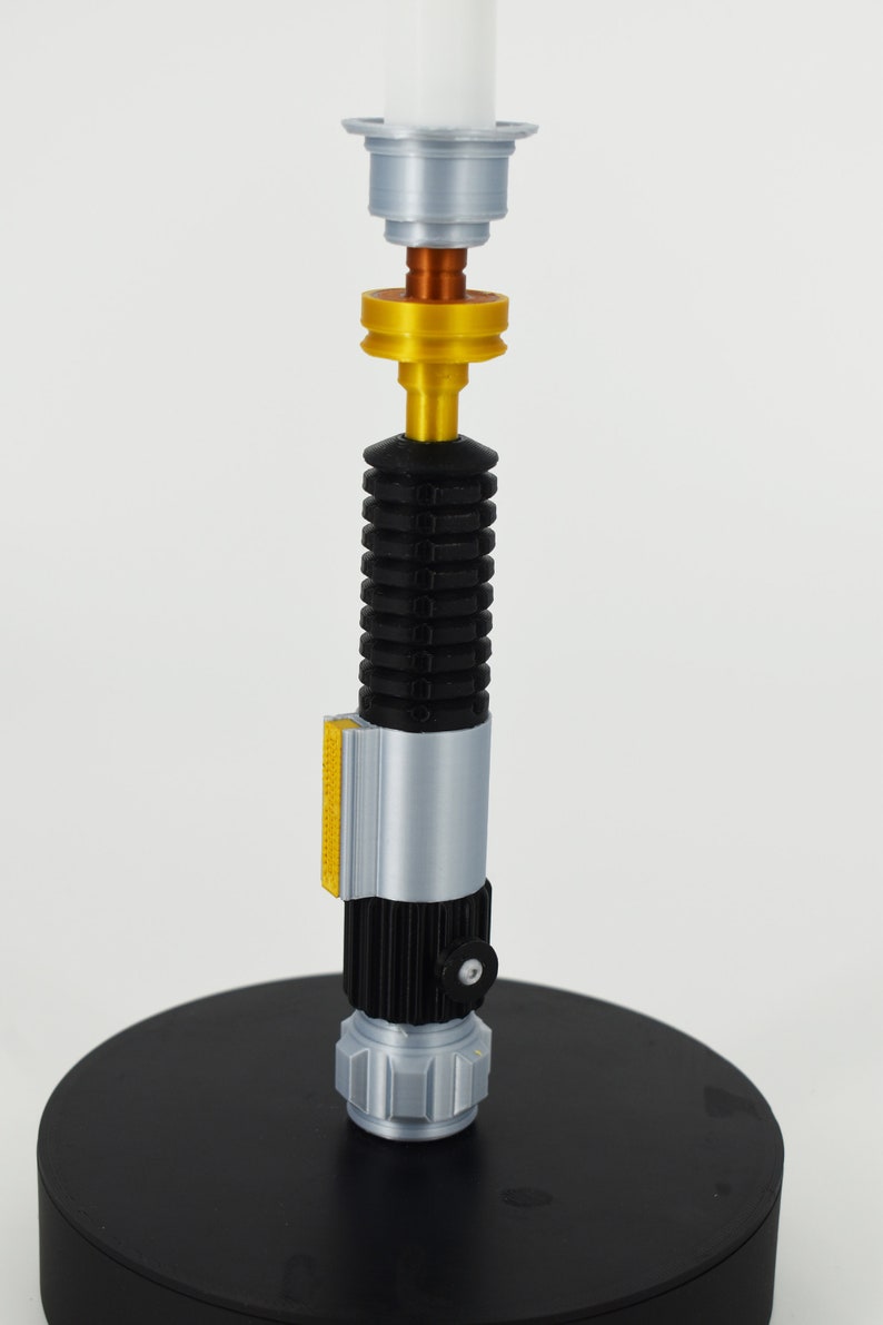 K&ouml;nnte beinhalten: Eine silberne und schwarze Lichtschwert-Replik mit einem goldenen Akzentringe und einer wei&szlig;en Spitze. Das Lichtschwert steht auf einem schwarzen Sockel.