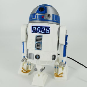 May include: A white and blue R2-D2 robot-shaped clock with a digital display showing the time. The clock has a silver dome and blue accents, with a small red light. The time displayed is 08:08.