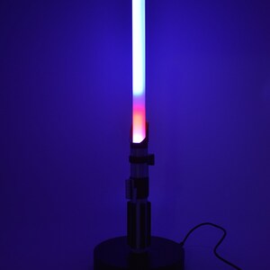 Op de afbeelding: Een zwarte lightsaber-lamp met een blauwe en rode lightsaber-blade. De lamp staat op een zwarte basis en heeft een snoer.