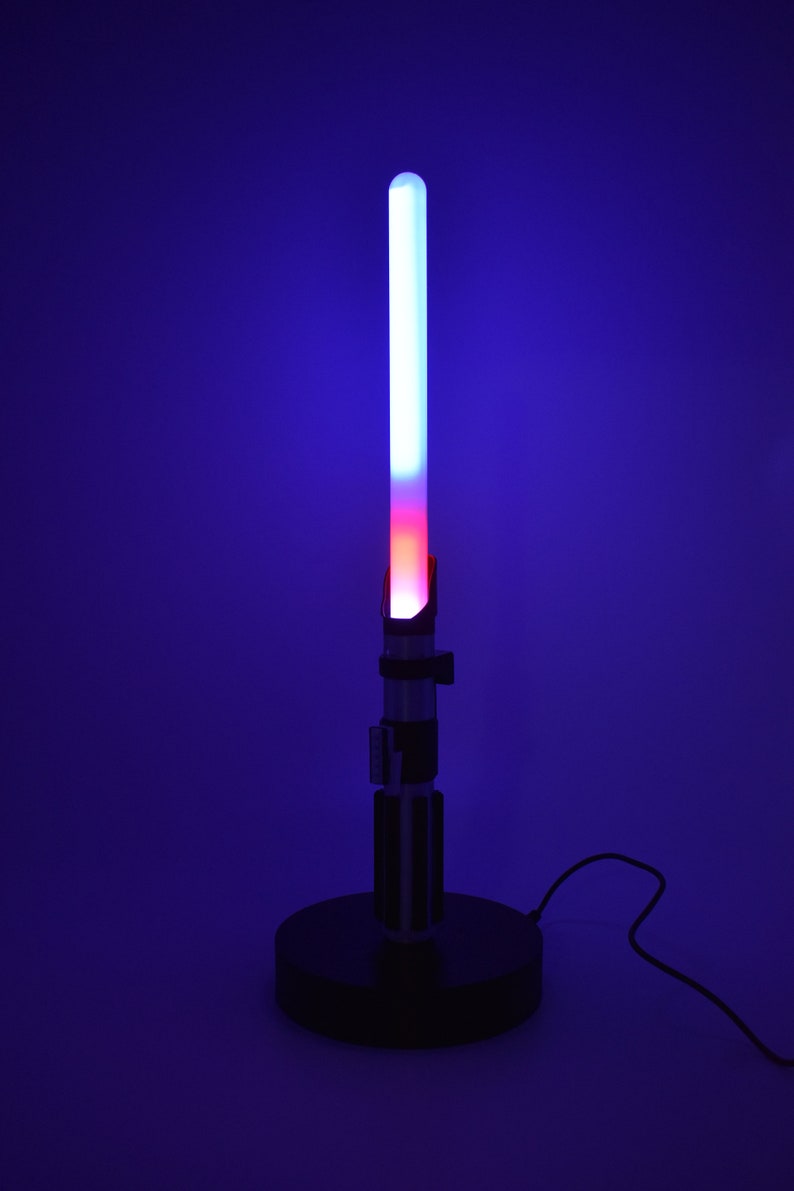 Op de afbeelding: Een zwarte lightsaber-lamp met een blauwe en rode lightsaber-blade. De lamp staat op een zwarte basis en heeft een snoer.
