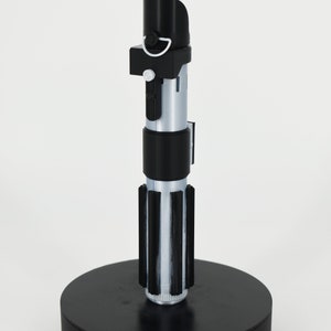Op de afbeelding: Een zwarte lightsaber-lamp met een witte blade en een zwarte voet. De voet heeft een aan/uit-schakelaar en een stroomkabel.