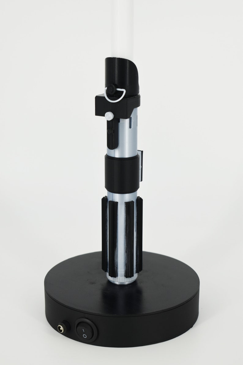 Op de afbeelding: Een zwarte lightsaber-lamp met een witte blade en een zwarte voet. De voet heeft een aan/uit-schakelaar en een stroomkabel.