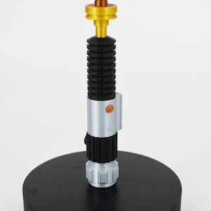 K&ouml;nnte beinhalten: Eine schwarze Lichtschwert-Lampe mit silbernem und goldenem Sockel. Die Lampe hat einen schwarzen Schalter am Sockel.