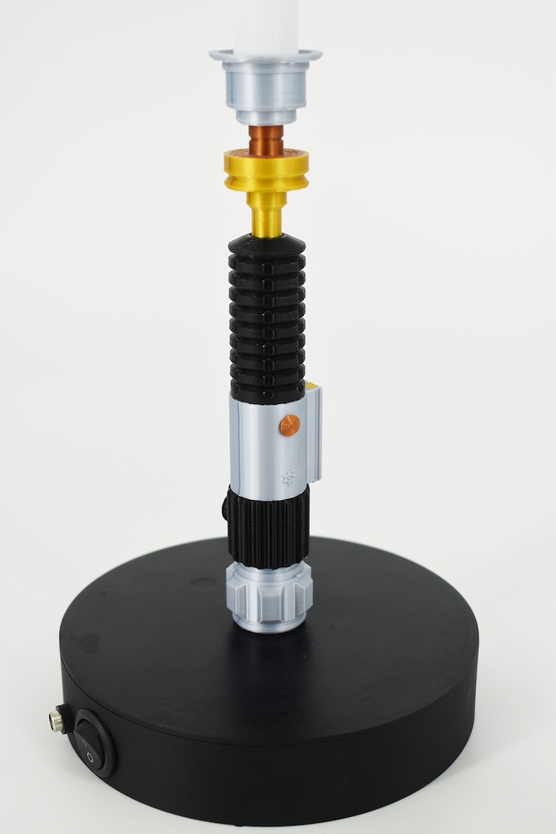 K&ouml;nnte beinhalten: Eine schwarze Lichtschwert-Lampe mit silbernem und goldenem Sockel. Die Lampe hat einen schwarzen Schalter am Sockel.