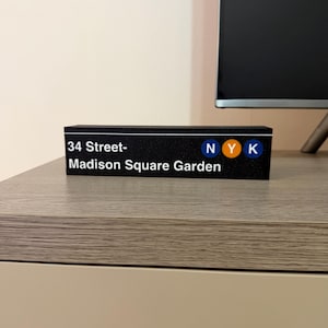 以下が含まれることがあります： 黒い長方形の看板で、白で「34 Street-Madison Square Garden」と書かれています。看板には青、オレンジ、青で「NYK」の文字も表示されています。看板は明るい色の表面に置かれています。