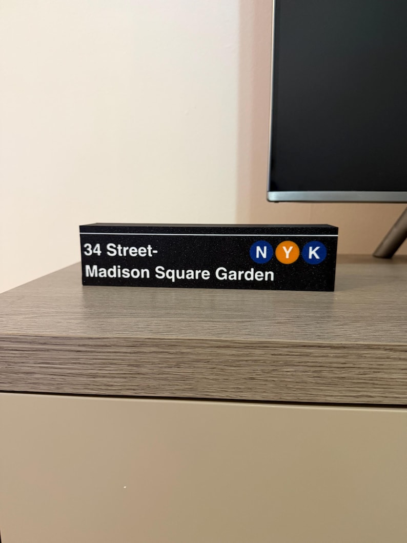 May include: A black rectangular sign with the text "34 Street-Madison Square Garden" in white. The sign also features the letters "NYK" in blue, orange, and blue. The sign is sitting on a light-colored surface.