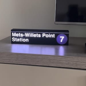 May include: A black sign with white text reading "Mets-Willetts Point Station" and a purple circle with the number "7". The sign is on a light-colored surface.