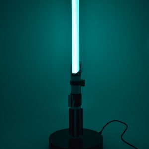 Op de afbeelding: Een zwarte lightsaber-lamp met een blauwe lightsaber-blade. De lamp staat op een zwarte basis en heeft een snoer.