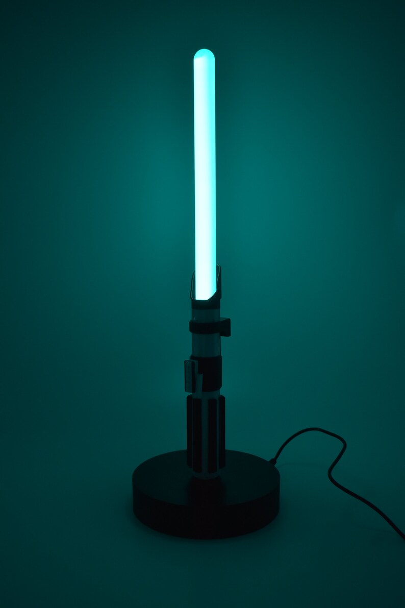 Op de afbeelding: Een zwarte lightsaber-lamp met een blauwe lightsaber-blade. De lamp staat op een zwarte basis en heeft een snoer.