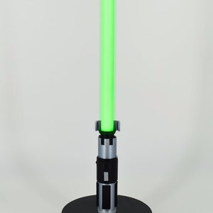 K&ouml;nnte beinhalten: Ein gr&uuml;nes Lichtschwert mit silbernem und schwarzem Griff, stehend auf einem schwarzen, kreisf&ouml;rmigen Sockel. Die Klinge des Lichtschwertes leuchtet hellgr&uuml;n. Der Griff hat detaillierte silberne und schwarze Akzente.