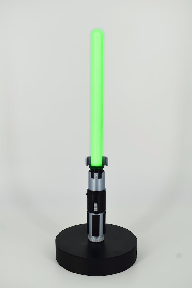 K&ouml;nnte beinhalten: Ein gr&uuml;nes Lichtschwert mit silbernem und schwarzem Griff, stehend auf einem schwarzen, kreisf&ouml;rmigen Sockel. Die Klinge des Lichtschwertes leuchtet hellgr&uuml;n. Der Griff hat detaillierte silberne und schwarze Akzente.