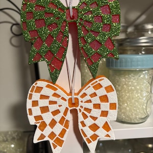 May include: Two decorative bow ornaments. The top bow has a red and green glitter design, with a diamond pattern. The bottom bow is orange with a white checkered pattern. Both bows have a string for hanging.