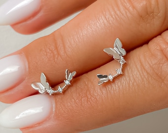 May include: A pair of silver-toned ear climbers featuring a curved design with two butterfly motifs and two small clear stones set between them. The earrings follow the natural curve of the earlobe, showcasing a polished metallic finish.