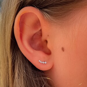 May include: A close-up of a person's ear with a small, silver ear cuff with five clear stones. The ear cuff is worn in the upper helix piercing.