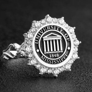 Könnte beinhalten: Ein silberner Ring mit dem Emblem der University of Mississippi. Das runde Gesicht des Rings zeigt das Universitätslogo und das Jahr 1848. Das Gesicht ist von einem Kranz aus kleinen, klaren Edelsteinen umgeben.