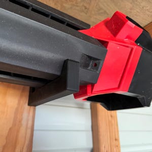 May include: A black and red tool component, mounted on wooden supports. The component has a rectangular shape with a series of vents and a red accent. The image is taken from a low angle, highlighting the design and construction.