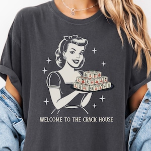 May include: Dark grey t-shirt featuring a retro illustration of a woman holding a tray of game tiles. The text "WELCOME TO THE CRACK HOUSE" is printed below the image. The shirt has short sleeves and a relaxed fit.