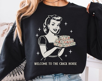 May include: Black long-sleeve sweatshirt featuring a retro illustration of a woman holding a tray of game tiles. The text "WELCOME TO THE CRACK HOUSE" is printed below the graphic. The sweatshirt is worn by a person.