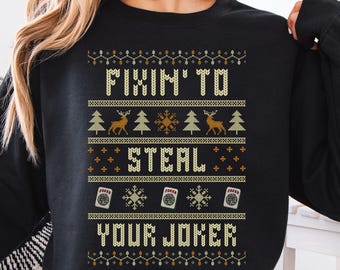 May include: A black long-sleeve sweatshirt featuring a graphic print in yellow, white, and orange. The design includes the text "FIXIN' TO STEAL YOUR JOKER" surrounded by patterns of reindeer, pine trees, snowflakes, and playing card icons.