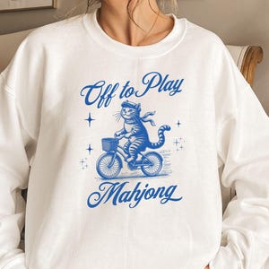 May include: White sweatshirt with a blue illustration of a cat riding a bicycle with a basket. The text "Off to Play Mahjong" is written above and below the cat in blue script.