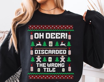 May include: A black long-sleeve sweatshirt featuring a festive graphic print with the text "OH DEER! DISCARDED THE WRONG TILE". The design includes red and green patterns, reindeer, gingerbread figures, and mahjong tiles.