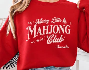 May include: A red long-sleeve crewneck sweatshirt featuring white text that reads Merry Little MAHJONG Club with a small Christmas tree and a silhouette of Santa in his sleigh, finished with the name Amanda printed below the text.