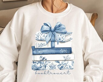 May include: White sweatshirt featuring a stack of books with blue floral designs and a large blue bow. The word "booktrovert" is written in a script font below the books. A cozy, book-themed design.