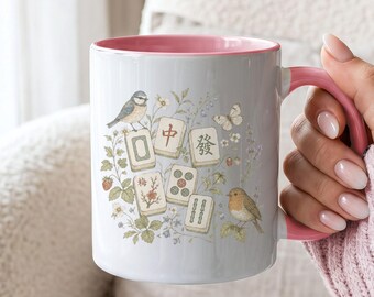 May include: A white ceramic mug with a pink interior and handle, featuring a botanical illustration of Mahjong tiles, small birds, butterflies, and strawberries. The design includes delicate floral vines and greenery surrounding the tiles.