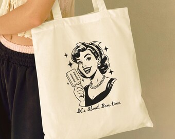 May include: A white canvas tote bag featuring a black graphic print of a smiling person holding a popsicle. The design includes four-pointed star accents and the text "It's About Bam time" written in a cursive script font below the illustration.