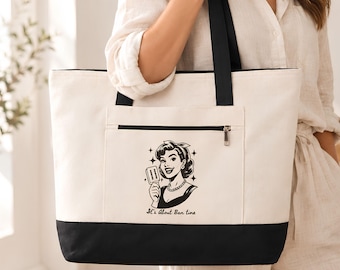 May include: A cream-colored canvas tote bag with black straps and a black base. The front features a zippered pocket with a black graphic illustration of a person holding a popsicle and the text "It's About Ban tine" printed below.