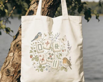 May include: A cream-colored canvas tote bag featuring a central illustration of five mahjong tiles surrounded by botanical elements, a blue tit, a robin, and a butterfly. The tiles display red and green Chinese characters and symbols.