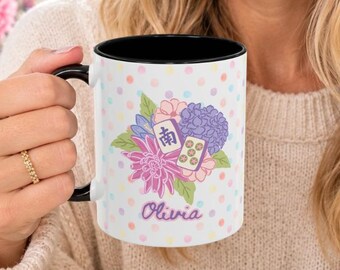 May include: A white ceramic mug with a black interior and handle, featuring a colorful polka dot pattern, a floral arrangement, two mahjong tiles, and the name Olivia printed in purple script on the front surface of the mug.