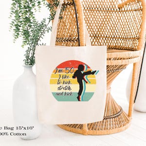 Puede incluir: Una bolsa de tela de algod&oacute;n de color natural que mide 38 cm por 41 cm. El frente presenta un gr&aacute;fico circular con un dise&ntilde;o de atardecer retro en rojo, amarillo y verde azulado, mostrando la silueta de una persona pateando y el texto I'm 50 I like to kick. stretch. and kick.