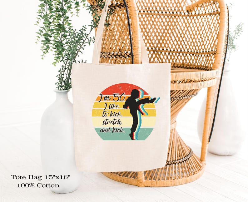 Puede incluir: Una bolsa de tela de algod&oacute;n de color natural que mide 38 cm por 41 cm. El frente presenta un gr&aacute;fico circular con un dise&ntilde;o de atardecer retro en rojo, amarillo y verde azulado, mostrando la silueta de una persona pateando y el texto I'm 50 I like to kick. stretch. and kick.