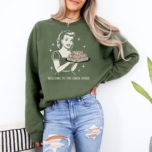 Op de afbeelding: Olijfgroene sweatshirt met een retro afbeelding van een vrouw met een dienblad met lekkernijen. De tekst "WELCOME TO THE CRACK HOUSE" is inbegrepen. De persoon draagt een blauwe jeans.