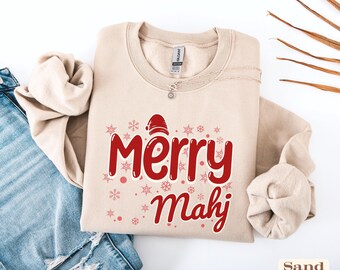 May include: A sand-colored crewneck sweatshirt featuring the red text Merry Mahj with a small red Santa hat on the letter M. The design is surrounded by various red snowflake icons and small dots, printed on the front of the garment.