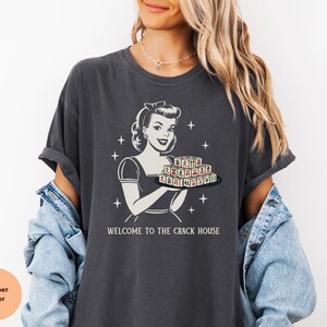 Op de afbeelding: Donkergrijze t-shirt met een retro-illustratie van een vrouw die een dienblad met speltegels vasthoudt. De tekst "WELCOME TO THE CRACK HOUSE" staat eronder. Het t-shirt heeft korte mouwen en een relaxte pasvorm.