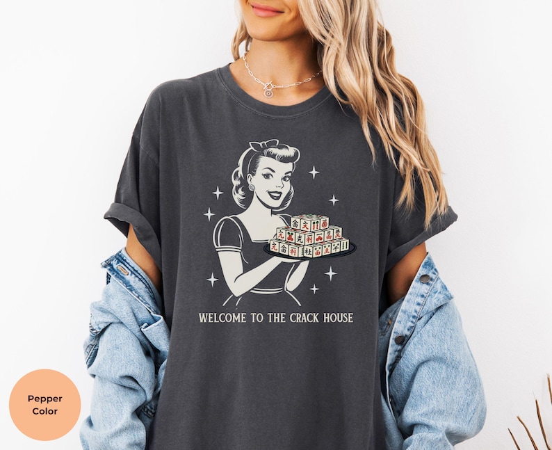 Op de afbeelding: Donkergrijze t-shirt met een retro-illustratie van een vrouw die een dienblad met speltegels vasthoudt. De tekst "WELCOME TO THE CRACK HOUSE" staat eronder. Het t-shirt heeft korte mouwen en een relaxte pasvorm.