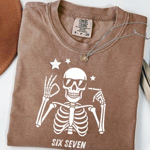 以下が含まれることがあります： サングラスをかけた白い骸骨のグラフィックが特徴の茶色のTシャツ。骸骨の下には「SIX SEVEN」の文字。ペンダント付きのシルバーネックレスがシャツの上にあります。