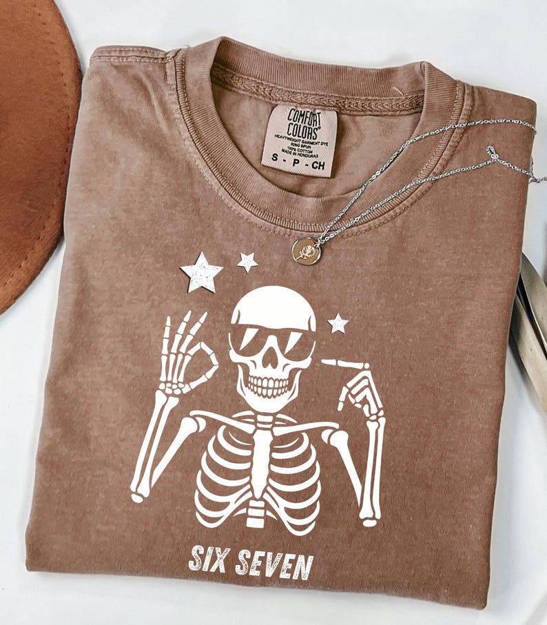 以下が含まれることがあります： サングラスをかけた白い骸骨のグラフィックが特徴の茶色のTシャツ。骸骨の下には「SIX SEVEN」の文字。ペンダント付きのシルバーネックレスがシャツの上にあります。