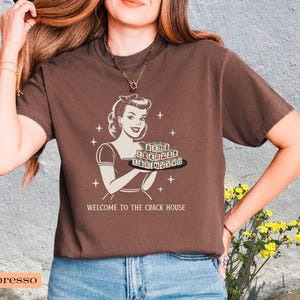 Op de afbeelding: Espresso-kleurig t-shirt met een retro-illustratie van een vrouw die een dienblad vasthoudt, met de tekst "WELCOME TO THE CRACK HOUSE". Het ontwerp is in vintage stijl, met steraccenten.