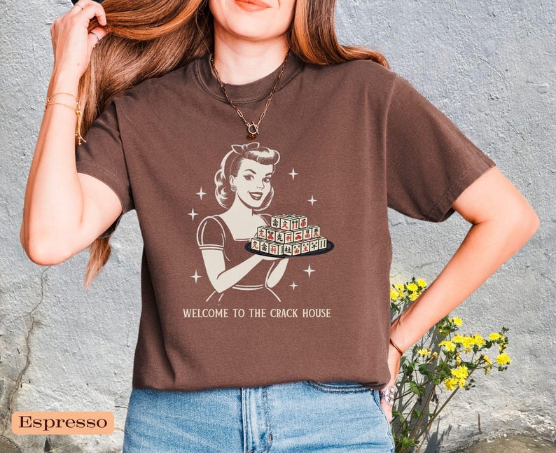 Op de afbeelding: Espresso-kleurig t-shirt met een retro-illustratie van een vrouw die een dienblad vasthoudt, met de tekst "WELCOME TO THE CRACK HOUSE". Het ontwerp is in vintage stijl, met steraccenten.
