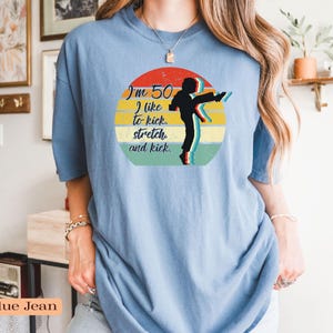 Puede incluir: Una camiseta de color azul vaquero con un gr&aacute;fico circular que muestra un atardecer retro en rojo, amarillo y verde. El dise&ntilde;o incluye la silueta negra de una persona dando una patada y el texto: I'm 50. I like to kick, stretch, and kick.