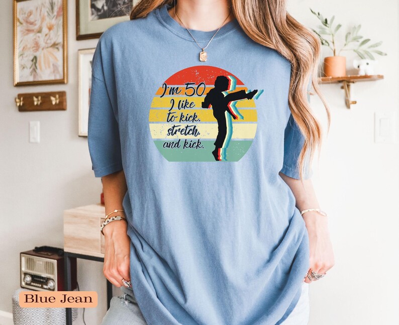 Puede incluir: Una camiseta de color azul vaquero con un gr&aacute;fico circular que muestra un atardecer retro en rojo, amarillo y verde. El dise&ntilde;o incluye la silueta negra de una persona dando una patada y el texto: I'm 50. I like to kick, stretch, and kick.